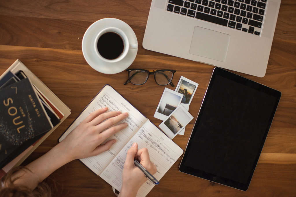 Person holding ballpoint pen writing on a notebook on a busy desk.