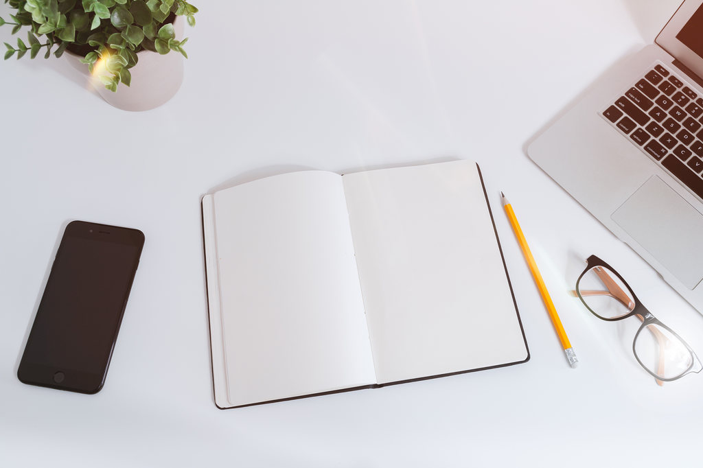 White desk with an open, empty notebook, pen, smartphone, and laptop.