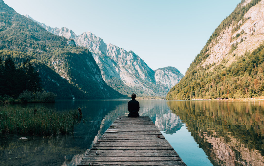 A person sitting on a dock of a beautiful lake surrounded by mesmerizing mountains.