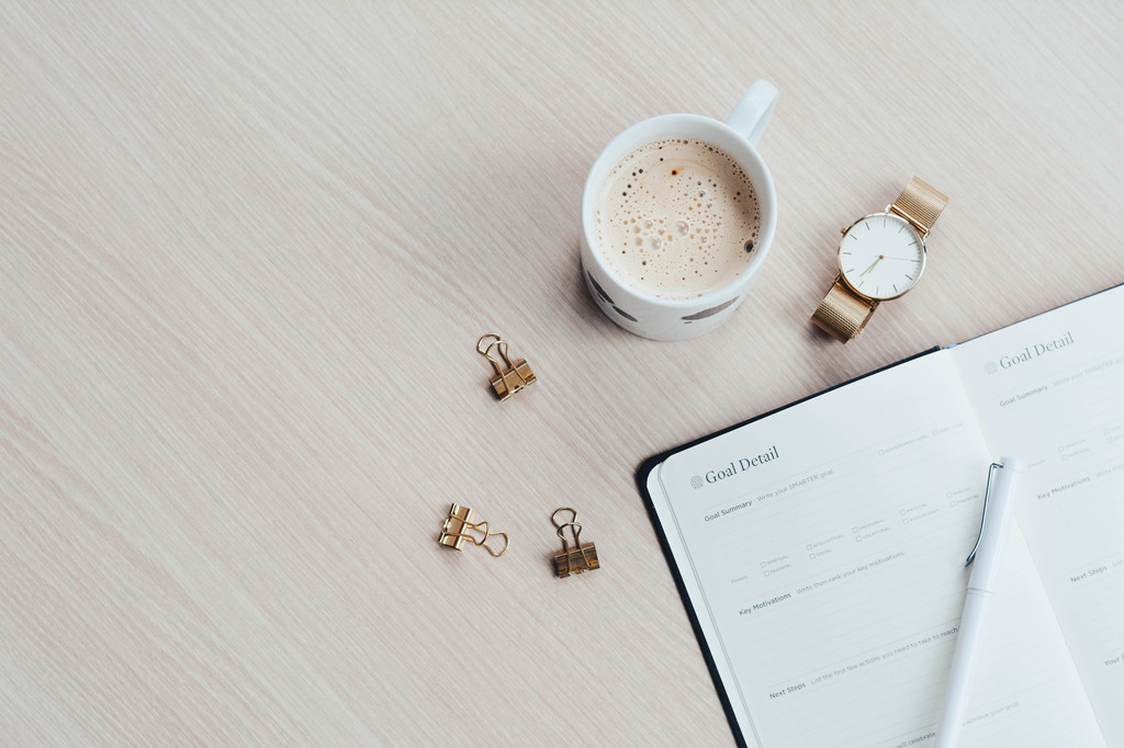 An open journal on a desk beside a filled mug with the title "Goal Detail" on top.