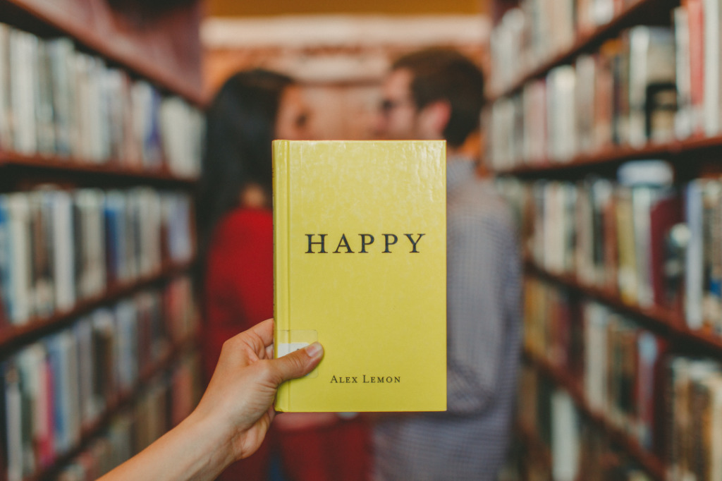 A person holding a book Happy by Alex Lemon, with a couple in the background holding hands like they're about to kiss.