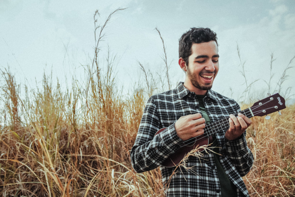 A man playing ukulele and smiling while standing on grass field.