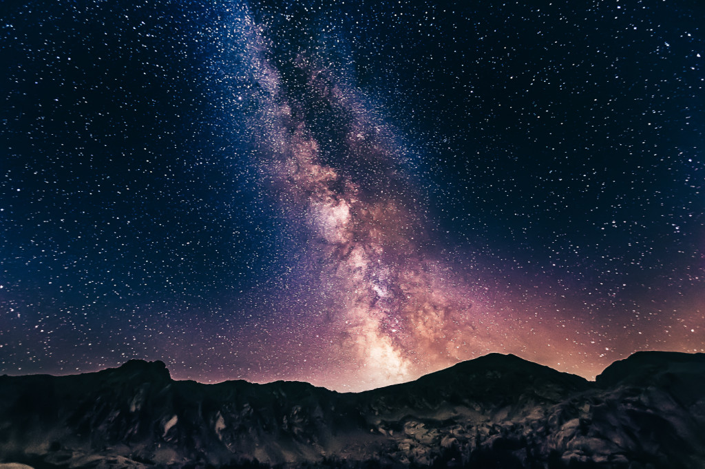 A night sky full of stars with a Milky Way galaxy in the center, behind mountains.