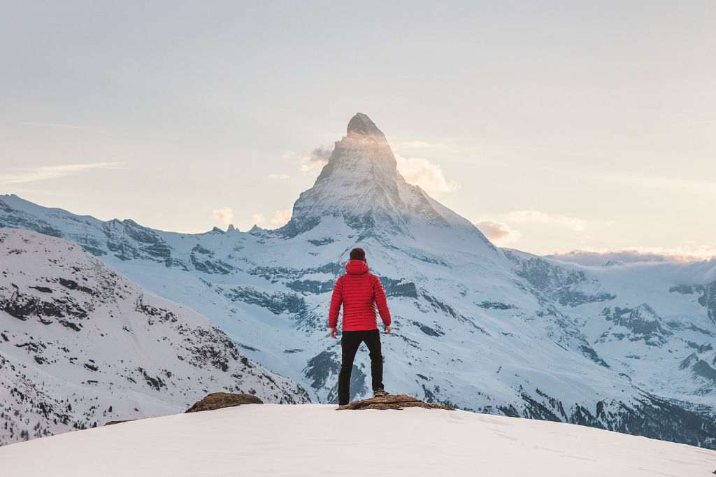 A person in a red hoodie standing on snow, looking at a distant mountain top.